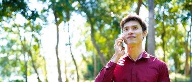 Parkta yürürken akıllı telefon kullanan mutlu Asyalı bir adam güneşli bir günde, yemyeşil ve modern mimariyle çevrili güzel bir sohbetin tadını çıkarıyor..