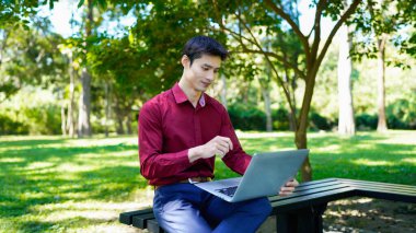 Parktaki bankta oturan bir adam yanında kahve fincanıyla dizüstü bilgisayar kullanarak güneşli bir günün tadını çıkarıyor..