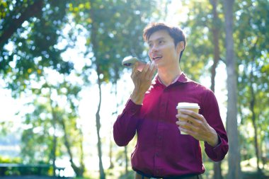 Parkta akıllı telefon kullanan mutlu Asyalı bir adam, güneşli bir günde keyifli bir sohbetin tadını çıkarıyor, yemyeşil ve modern mimariyle çevrili...