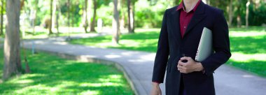 Siyah takım elbiseli profesyonel Asyalı işadamı parkta dizüstü bilgisayar taşıyor..