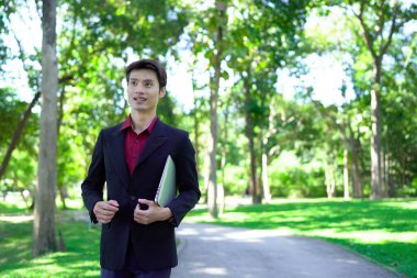 Siyah takım elbiseli profesyonel Asyalı işadamı parkta dizüstü bilgisayar taşıyor..