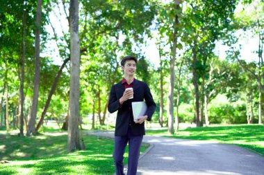 Siyah takım elbiseli profesyonel Asyalı işadamı elinde dizüstü bilgisayarla parkta kahve fincanı tutuyor...