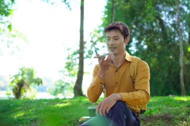 Parkta akıllı telefon kullanan mutlu Asyalı bir adam, güneşli bir günde keyifli bir sohbetin tadını çıkarıyor, yemyeşil ve modern mimariyle çevrili...