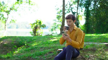 Paket kahveli genç Asyalı adam şehir parkında akıllı telefondan iletişim kuruyor..