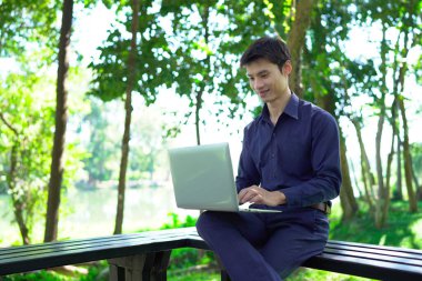 Genç adam güneşli bir park ortamında dizüstü bilgisayarda çalışırken aynı zamanda canlı ve profesyonel bir yaşam tarzının tadını çıkarıyor..