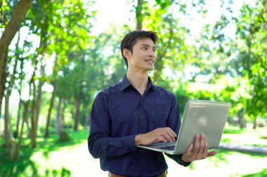 Genç adam güneşli bir park ortamında dizüstü bilgisayarda çalışırken aynı zamanda canlı ve profesyonel bir yaşam tarzının tadını çıkarıyor..