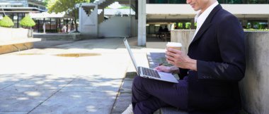 Parkta dizüstü bilgisayarda çalışan, parktaki bankta kahve içen genç bir adam..