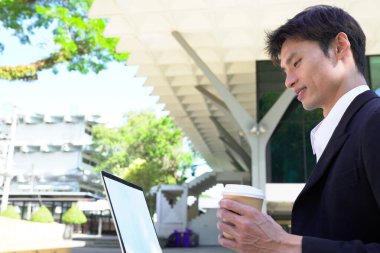 Parkta dizüstü bilgisayarda çalışan, parktaki bankta kahve içen genç bir adam..
