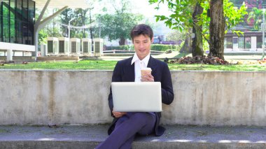 Parkta dizüstü bilgisayarda çalışan, parktaki bankta kahve içen genç bir adam..