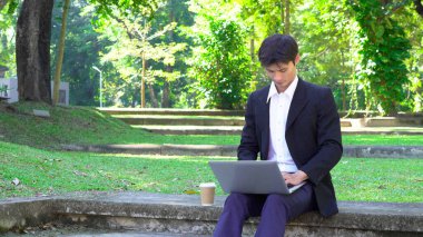 Parktaki bankta oturan bir adam yanında kahve fincanıyla dizüstü bilgisayar kullanarak güneşli bir günün tadını çıkarıyor..