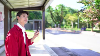 Akıllı telefonlu genç adam otobüs durağında toplu taşıma bekliyor..