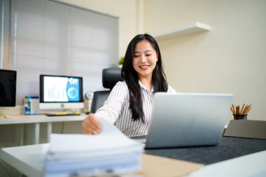 Dizüstü bilgisayarında çalışan iş kadını, masa başındaki yoğun işte, klasör kağıdındaki belgeleri aramak ve kontrol etmek için kağıt yığınıyla çalışıyor..