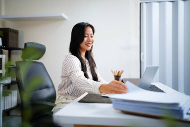 Dizüstü bilgisayarında çalışan iş kadını, masa başındaki yoğun işte, klasör kağıdındaki belgeleri aramak ve kontrol etmek için kağıt yığınıyla çalışıyor..