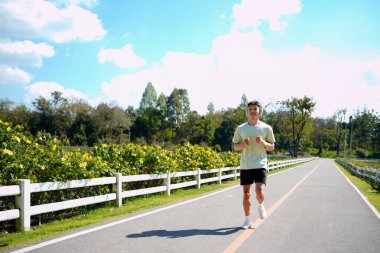 Asyalı bir adam parkta koşuyor, güneşli bir günde koşuyor, gülümsüyor ve açık havada eğleniyor...