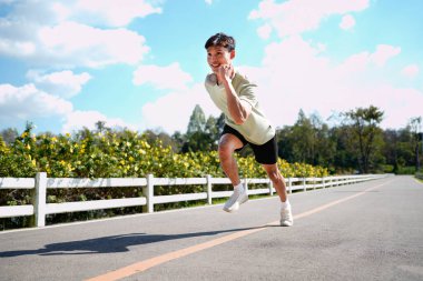 Asyalı bir adam parkta koşuyor, güneşli bir günde koşuyor, gülümsüyor ve açık havada eğleniyor...