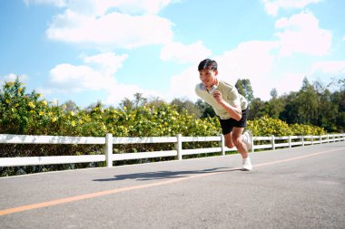 Asyalı bir adam parkta koşuyor, güneşli bir günde koşuyor, gülümsüyor ve açık havada eğleniyor...