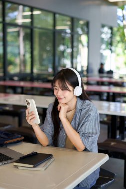Mutlu Asyalı öğrenci kampüste kulaklıkla müzik dinlerken cep telefonunu kullanıyor..