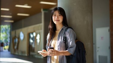 Genç Asyalı bir kadın işe giderken elinde kahve ve telefon çantası, sırtında sırt çantası şehrin çevresindeki kameralara bakar..