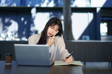 Asyalı serbest çalışan bir bayan öğrenci, telefonda konuşuyor. Dizüstü bilgisayarını bir kenara bırakıp binada oturuyor..