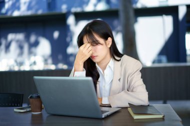 Bilgisayardaki bilgisayar ekranına odaklanmış genç bir iş kadınının baş ağrısı ve migreni var. Asyalı iş kadını şakakların yanında el ele tutuşuyor, stresli ve yorgun hissediyor ofis binasında oturuyor..