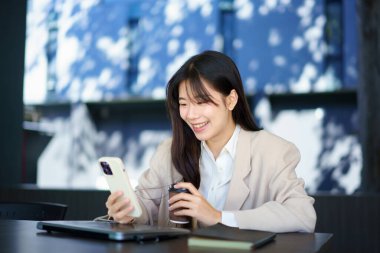 Asyalı kadın mutlu ve kafede akıllı telefondan mesaj yazıyor. Gülümseyen genç kadın sıcak bir fincan kahveyle masada oturuyor ve cep telefonu kullanıyor..