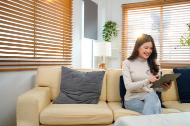 Modern beyaz oturma odasındaki koltukta oturan mutlu genç kadın, Tablet bilgisayarını kullanarak parlak doğal ışıkla çevrili içeriği görüyor. Güzel insanlar ve modern teknoloji konsepti...