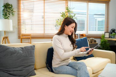 Modern beyaz oturma odasındaki koltukta oturan mutlu genç kadın, Tablet bilgisayarını kullanarak parlak doğal ışıkla çevrili içeriği görüyor. Güzel insanlar ve modern teknoloji konsepti...