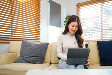 Modern beyaz oturma odasındaki koltukta oturan mutlu genç kadın, Tablet bilgisayarını kullanarak parlak doğal ışıkla çevrili içeriği görüyor. Güzel insanlar ve modern teknoloji konsepti...