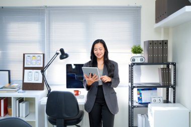 Güzel gülümseyen Asyalı iş kadını elinde tabletle ofiste çalışıyor ve iş detaylarını kaydediyor..