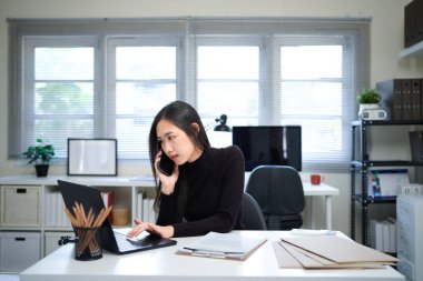 Genç Asyalı mutlu bir kadın cep telefonuyla konuşuyor ve ofisinde çalışırken gülümsüyor..