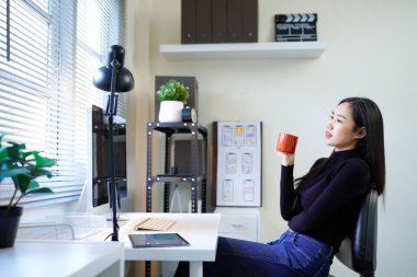 Güzel Asyalı kadın bilgisayar monitörü ve bir fincan kahveyle bir masada oturuyor..