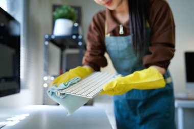 Genç Asyalı kadın, günlük hademe üniformalı, antiseptik sprey şişeli, ıslak bez temizleyici klavye bilgisayarı, masa ve masa yüzeyleri modern ofis odasının içinde..