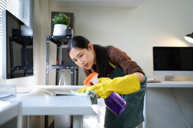 Genç Asyalı kadın, günlük hademe üniformalı, antiseptik sprey şişeli, ıslak bez temizleme masalı modern ofis odasının iç kısmında masa yüzeyleri olan hizmetli kadın..