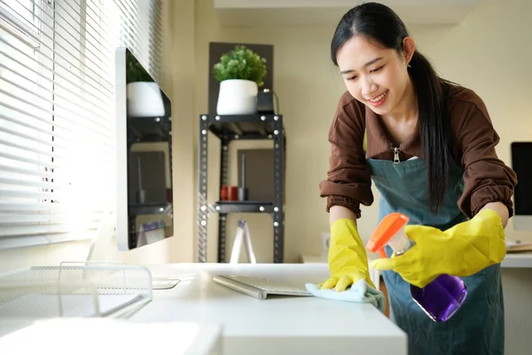 Genç Asyalı kadın, günlük hademe üniformalı, antiseptik sprey şişeli, ıslak bez temizleme masalı modern ofis odasının iç kısmında masa yüzeyleri olan hizmetli kadın..
