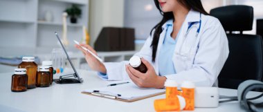 Kadın doktor ilaç şişesini incelerken modern klinikteki tablete notlar yazıyor..