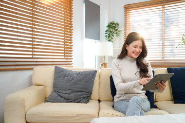 Modern beyaz oturma odasındaki koltukta oturan mutlu genç kadın, Tablet bilgisayarını kullanarak parlak doğal ışıkla çevrili içeriği görüyor. Güzel insanlar ve modern teknoloji konsepti..