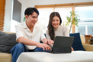 Çift evde kanepede oturup tablet oynuyor ve birlikte dinleniyorlar. Gülümsüyorlar ve mutlulukla vakit geçirmekten zevk alıyorlar..