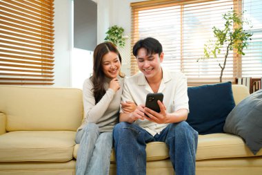 Mutlu genç kadın ve erkek sarılıyor, akıllı telefon kullanıyor, evdeki rahat koltukta oturuyor, aşırı mutlu bir şekilde gülümsüyor, telefon ekranına bakıyor, modern oturma odasındaki kanepede oturuyor..