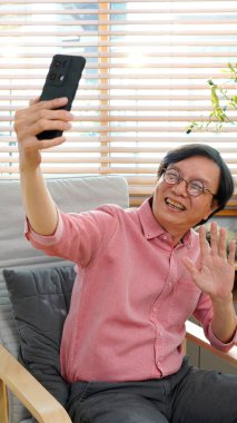 Akıllı telefonla selfie çekerken gülümseyen yaşlı bir adam. Bitkilerle ve yumuşak mobilyalarla dolu rahat bir oturma odası. Sosyal bağlantı kavramı, teknoloji.