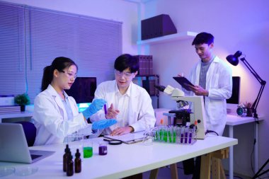 Bir araştırma laboratuvarında eldiven içinde çözeltisi olan bir test tüpü tutan Asyalı kadın bilim adamı veya laboratuvar önlüklü genç bilim adamları yüksek teknolojili bir laboratuvarda işbirliği yapıyorlar..