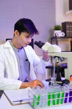 Asyalı erkek bilim adamı modern bir laboratuvarda mikroskop kullanarak araştırma yapıyor. Odaklanmış ve laboratuvar önlüğü giymiş..