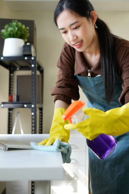 Genç Asyalı kadın, günlük hademe üniformalı, antiseptik sprey şişeli, ıslak bez temizleme masalı modern ofis odasının iç kısmında masa yüzeyleri olan hizmetli kadın..
