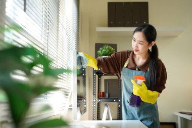 Genç Asyalı kadın, üniformalı hademe kadın antiseptik sprey şişesi ve ıslak bez temizleme masaüstü bilgisayarı, masa ve masa yüzeyleri modern ofis odasının içinde..