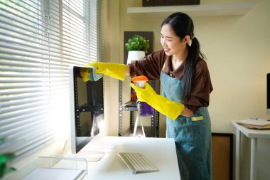 Genç Asyalı kadın, üniformalı hademe kadın antiseptik sprey şişesi ve ıslak bez temizleme masaüstü bilgisayarı, masa ve masa yüzeyleri modern ofis odasının içinde..