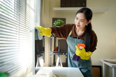 Genç Asyalı kadın, üniformalı hademe kadın antiseptik sprey şişesi ve ıslak bez temizleme masaüstü bilgisayarı, masa ve masa yüzeyleri modern ofis odasının içinde..