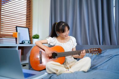 Bilgisayarla gitar çalan güzel bir genç kadın, online kursla oynamayı öğreniyor, evdeki gündelik kıyafetlerle kanepede oturuyor..