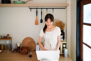 Mutfak tezgahında elinde kahveyle oturmuş gülümseyen Asyalı genç bir kadın ve bilgisayarını yemek tarifi aramak ya da küçük işletmelerini yönetmek için kullanıyor..