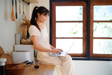 Mutfak tezgahında elinde kahveyle oturmuş gülümseyen Asyalı genç bir kadın ve bilgisayarını yemek tarifi aramak ya da küçük işletmelerini yönetmek için kullanıyor..