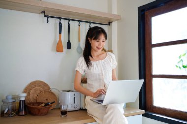 Mutfak tezgahında elinde kahveyle oturmuş gülümseyen Asyalı genç bir kadın ve bilgisayarını yemek tarifi aramak ya da küçük işletmelerini yönetmek için kullanıyor..