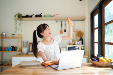 Asyalı kadın gülümsüyor, mutfak masasında dizüstü bilgisayarını yazarken bir fincan kahve ve ekmeğin keyfini çıkarıyor, uzaktan çalışma ve iş teknolojisini somutlaştırıyor..